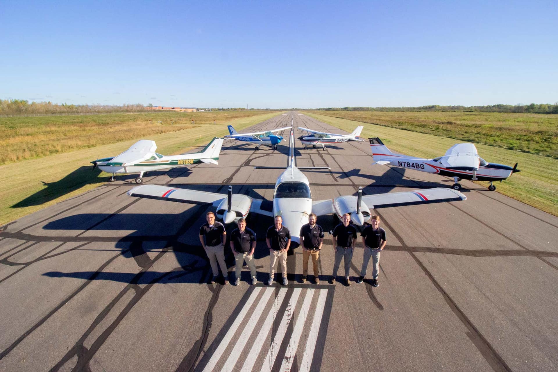Six members of the Superior Flying Services team standing in front of their twin-engine Piper Seneca on the runway at KSUW, with four Cessna 172s parked in a fan behind it.